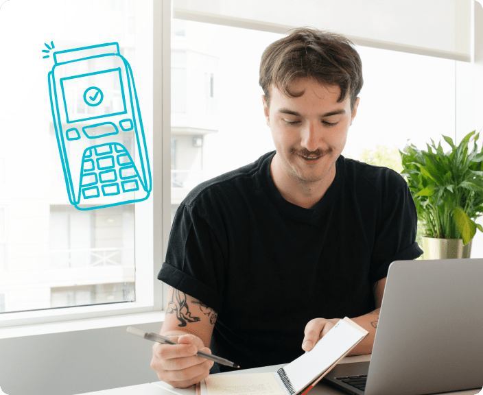 Young man at desk using laptop and smartphone with payment terminal icon indicating secure transaction processing
