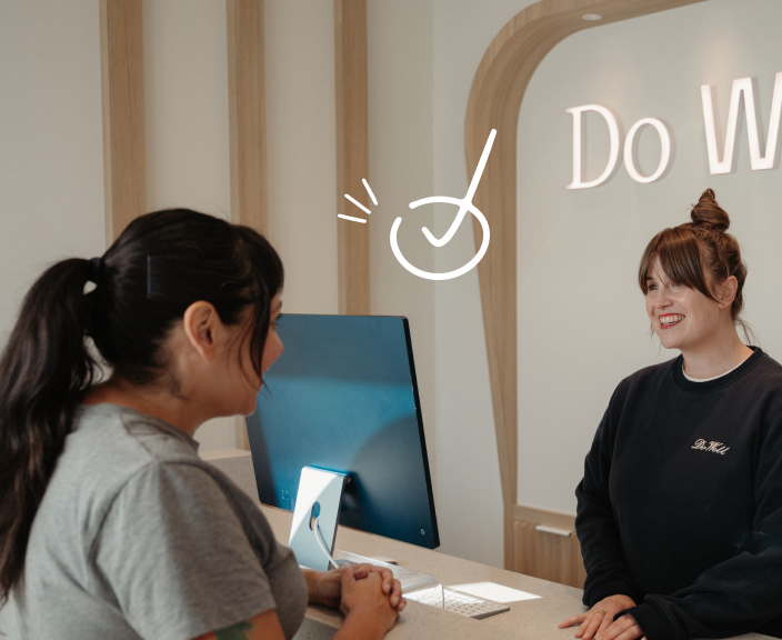 Two women at a reception desk, one seated at a computer helping a customer, with "Do We" text and checkmark icon visible on the wall behind them