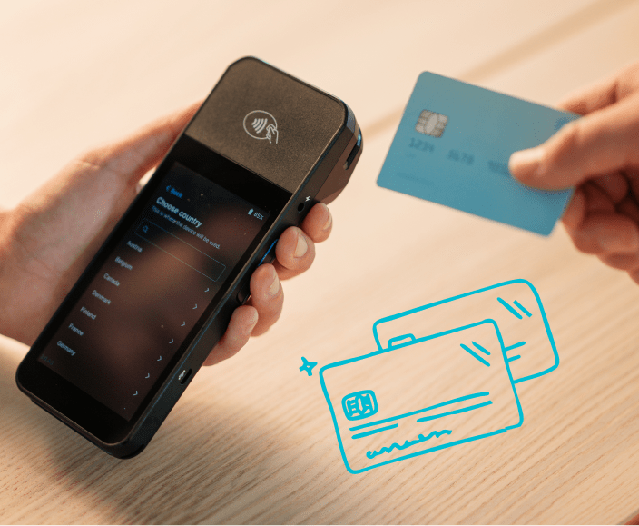 Hands holding a contactless payment terminal and blue credit card above illustrated payment cards on wooden surface