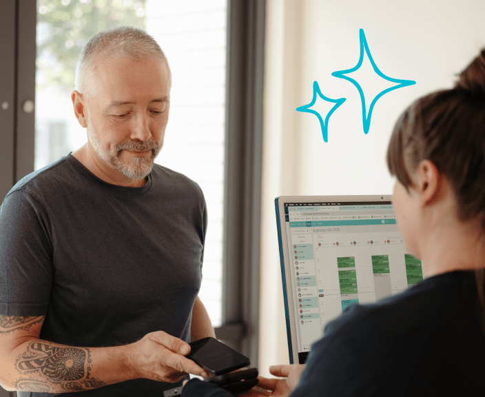 Man with gray beard and tattoo looking at phone while woman works at computer with scheduling software