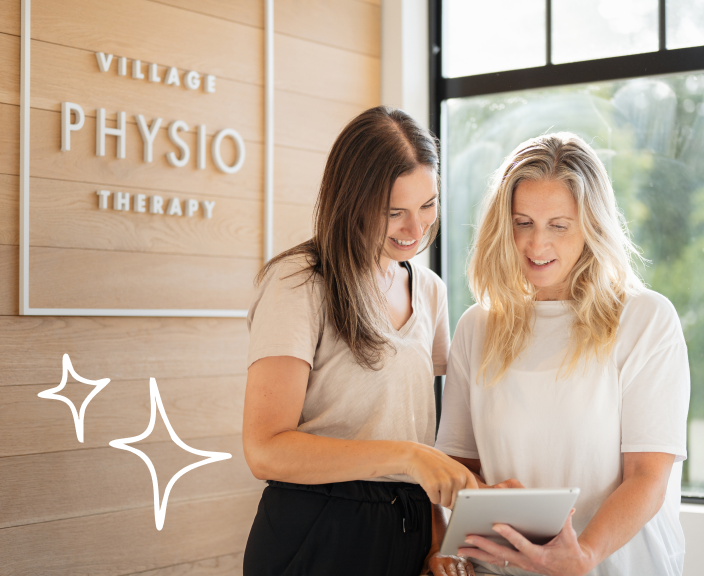 Two women in a physiotherapy clinic looking at a tablet together, with Village Physio Therapy signage visible on the wall