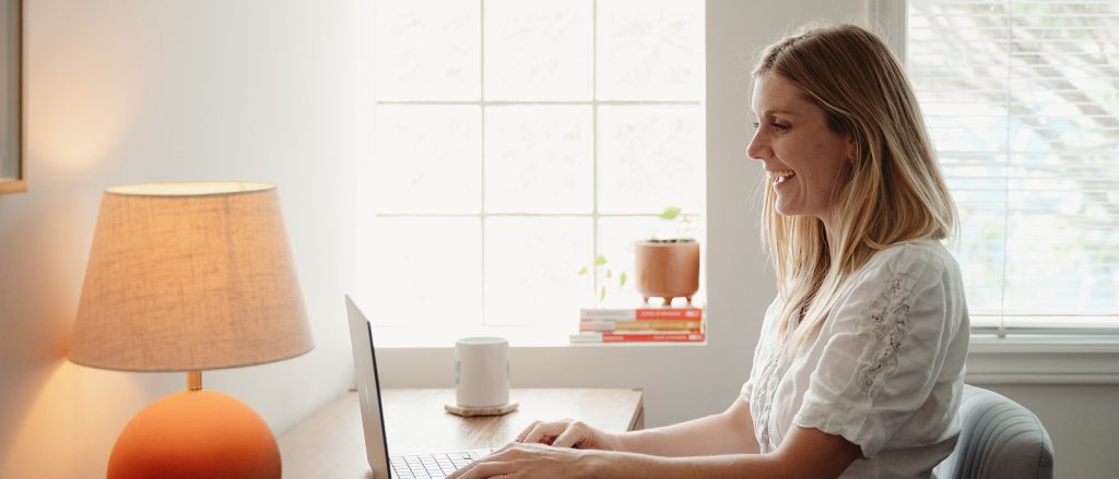 Woman working on laptop at desk by window in bright, naturally lit room