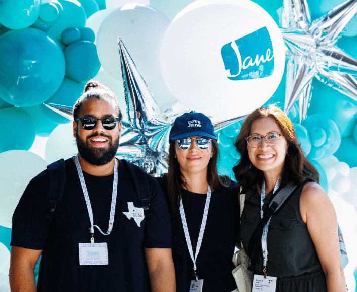 Three people wearing conference badges pose together in front of turquoise and silver balloon decorations with Jane branding