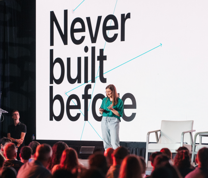 Speaker presenting on stage in front of an audience, with a screen behind reading "Never built before