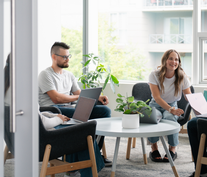Coworkers relax with laptops in a bright lounge area, chatting and smiling near large windows