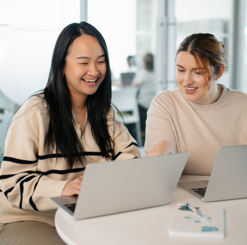 Two women smiling while collaborating on laptops together at a table in an office