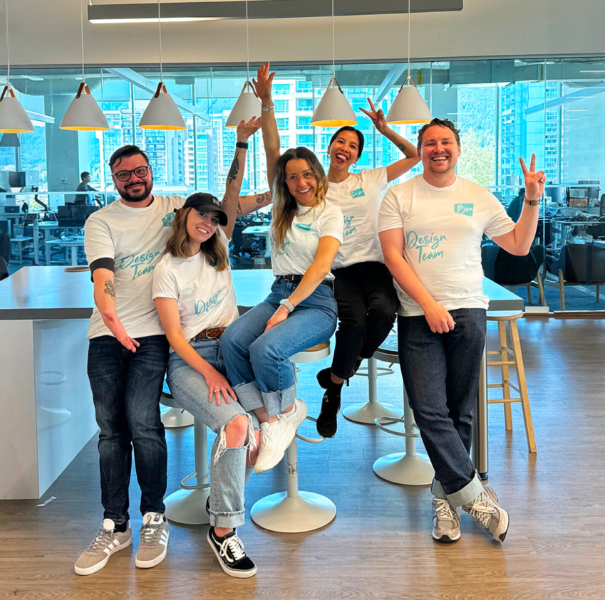 Five smiling team members in matching "Design Team" t-shirts pose playfully together in an office