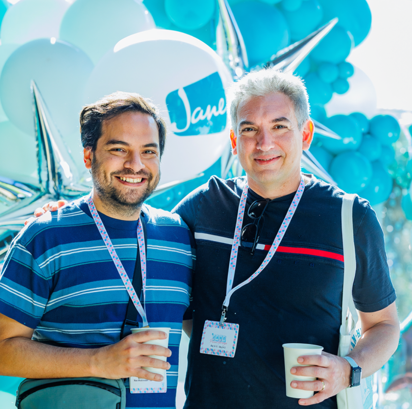 Two men wearing event lanyards smile together in front of teal and white balloon decorations at a Jane event