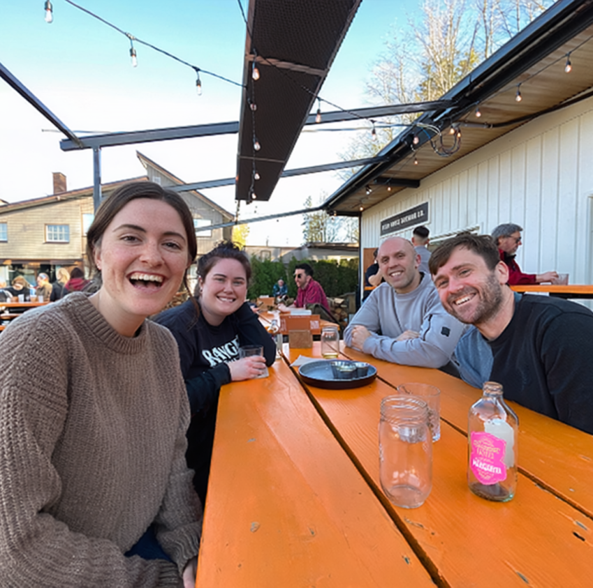 Four friends smiling around an outdoor picnic table with drinks, at a lively string-lit patio venue