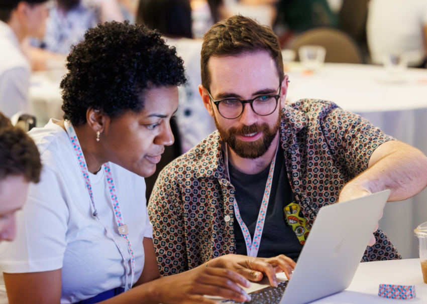 Two conference attendees wearing lanyards collaborate closely, looking at a laptop screen together