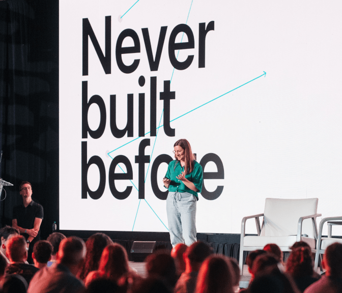 Speaker on stage in front of an audience, with a screen behind reading "Never built before