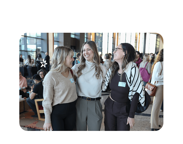 Three women wearing name badges laugh together at a crowded conference networking event